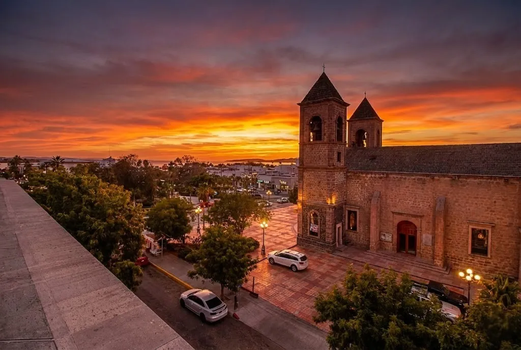 The historic twin-tower architecture of the Cathedral of Our Lady of La Paz facing the town square under a dramatic golden hour sunset in Baja California Sur.