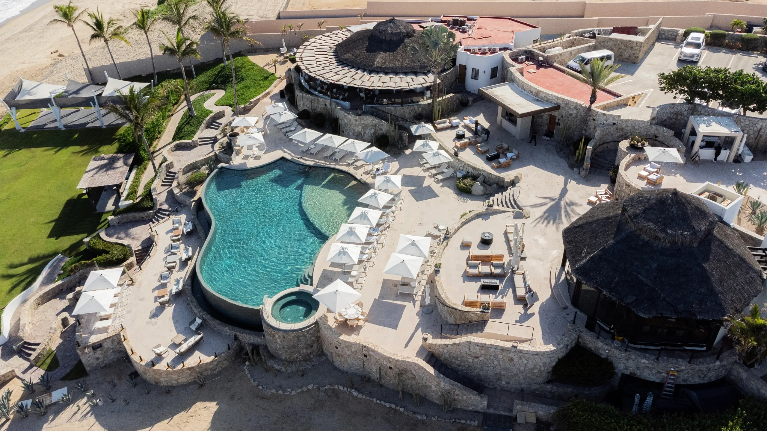High-angle aerial view of the exclusive Fundadores Beach Club in Puerto Los Cabos, featuring a large infinity pool, white lounging umbrellas, and oceanfront dining facilities. High-angle aerial view of the exclusive Fundadores Beach Club in Puerto Los Cabos, featuring a large infinity pool, white lounging umbrellas, and oceanfront dining facilities.