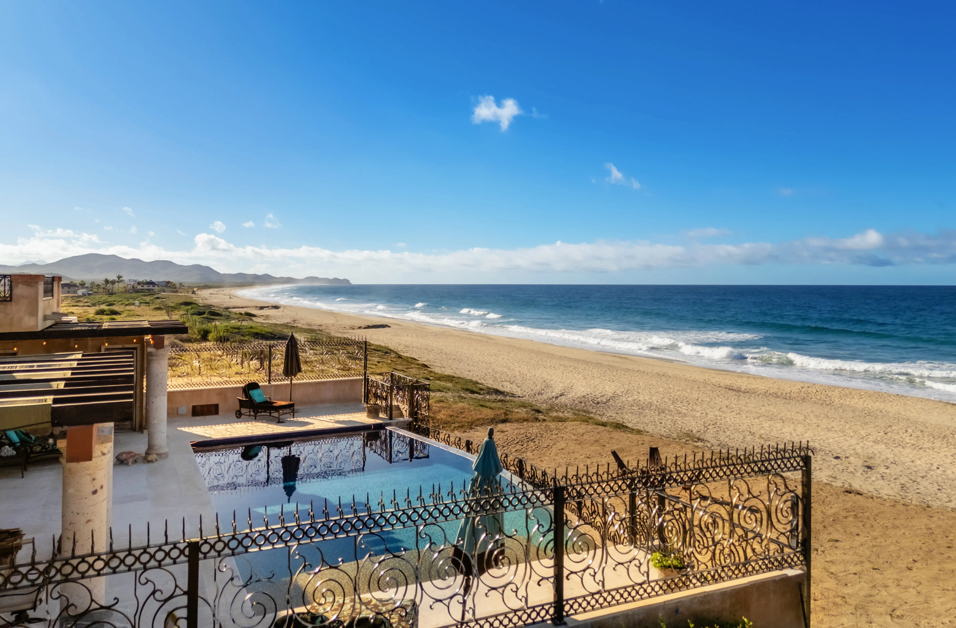 Aerial drone view of an oceanfront hacienda-style estate with infinity-edge pool, wrought iron railings, and pergola-covered terrace overlooking Cerritos Beach on the Baja California Sur Pacific coast.