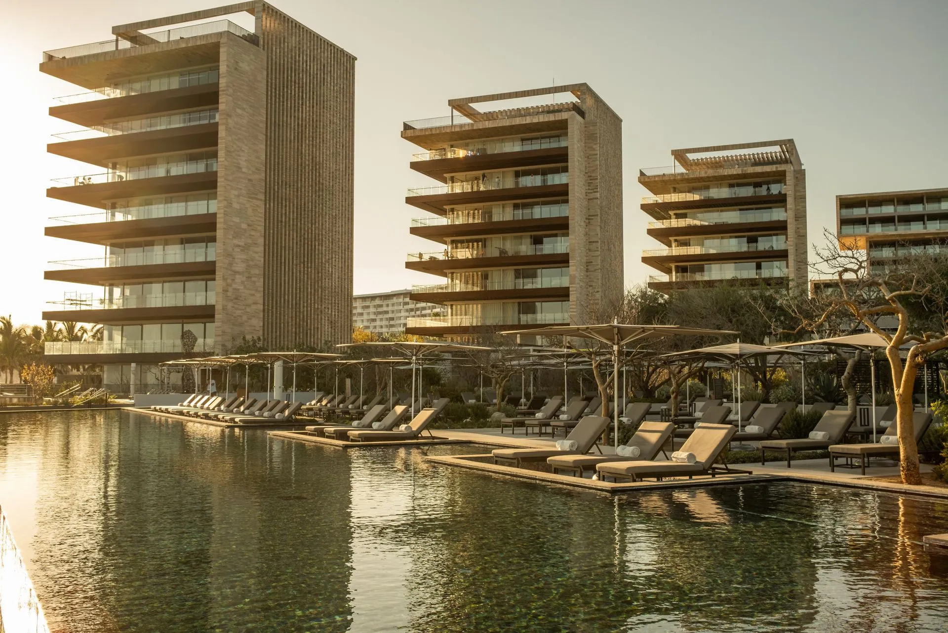 Main resort pool at Solaz Los Cabos at sunset, featuring luxury sun loungers, white umbrellas, and modern concrete-and-wood architecture bathed in golden hour light reflecting on the Sea of Cortés.