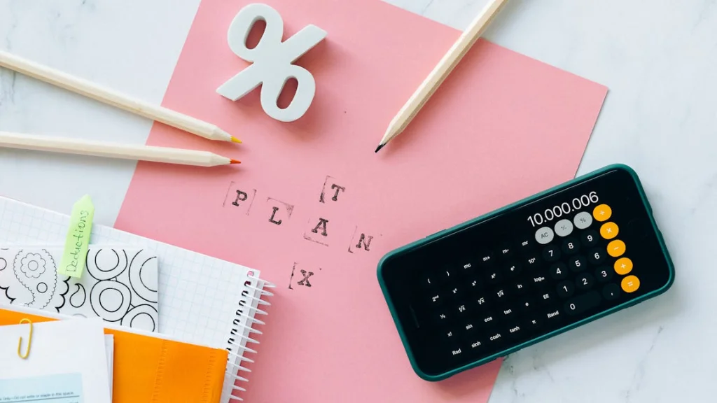 A pink sheet of paper with the words “tax and plan” printed on it, a cell phone with the calculator app open, three pencils, a notebook and a figure in the shape of a percent sign.